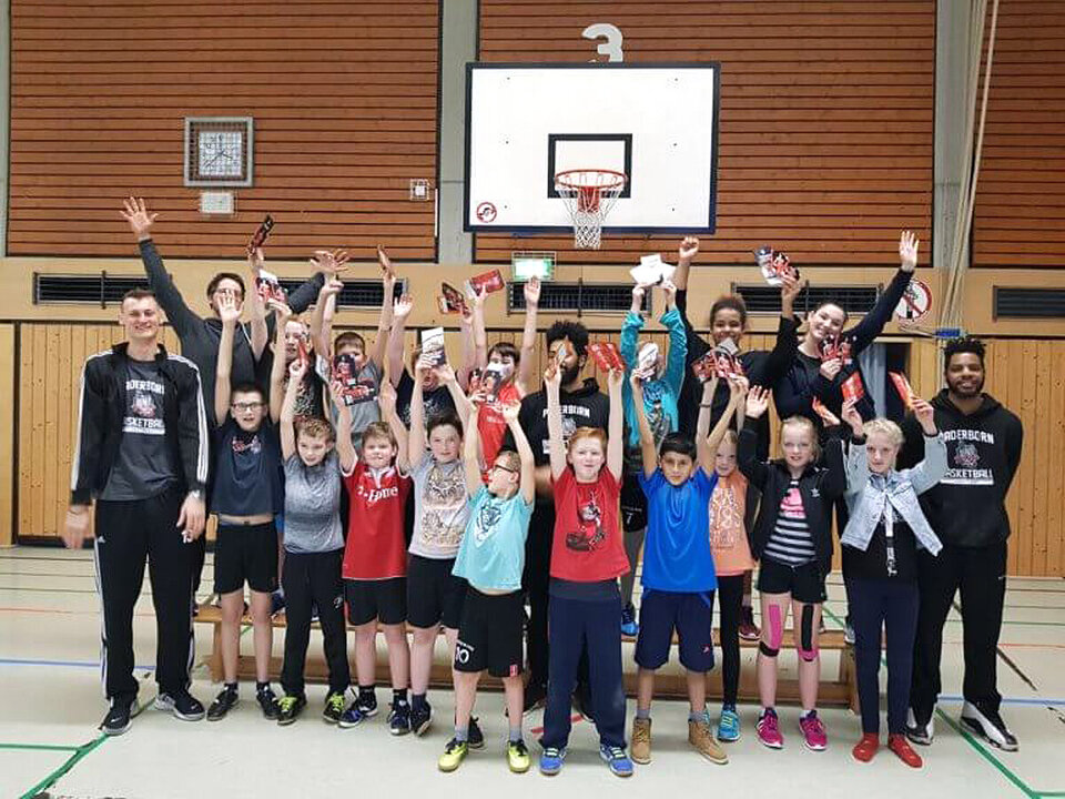 Eine Klasse steht zusammen unter einem Basketball-Korg und winkt in die Kamera